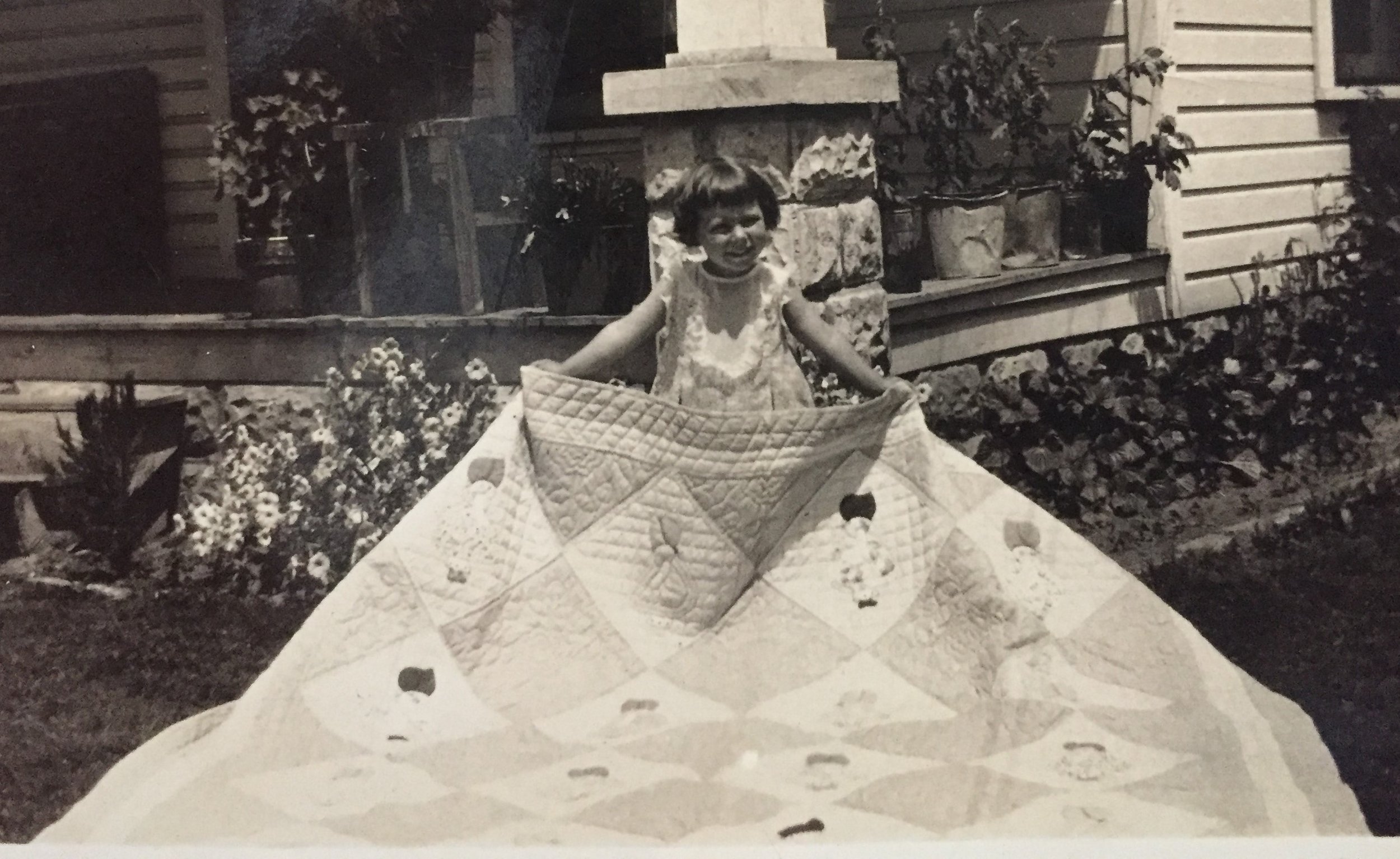 Vintage black-and-white photograph of a small girl holding up a large patchwork quilt.