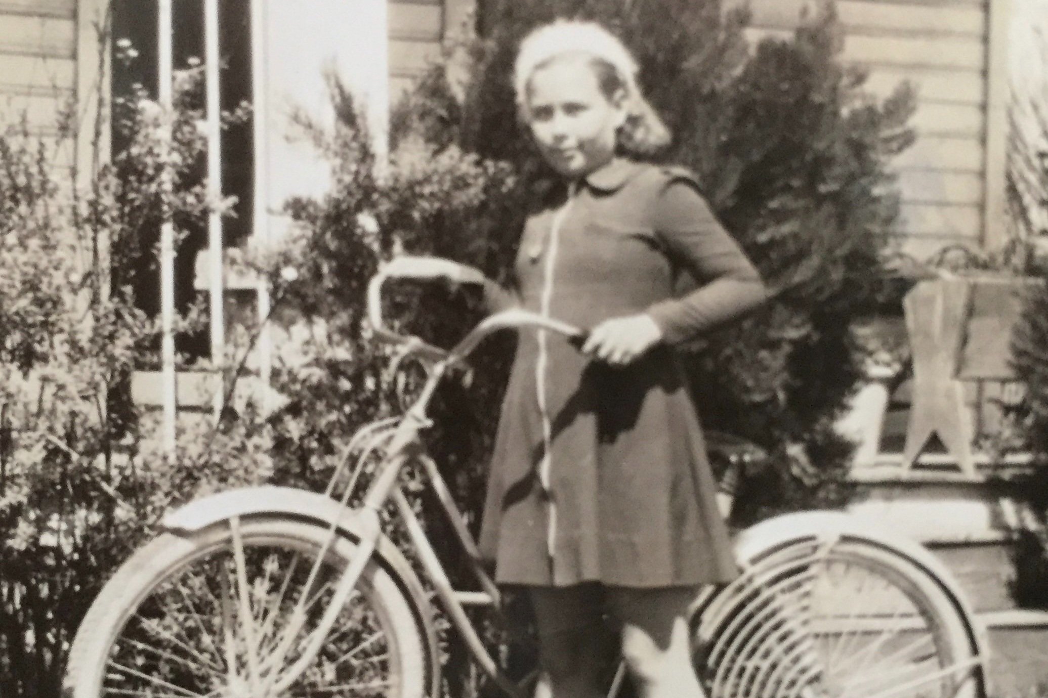 Vintage black-and-white photograph of a girl in a dress standing with a bicycle.