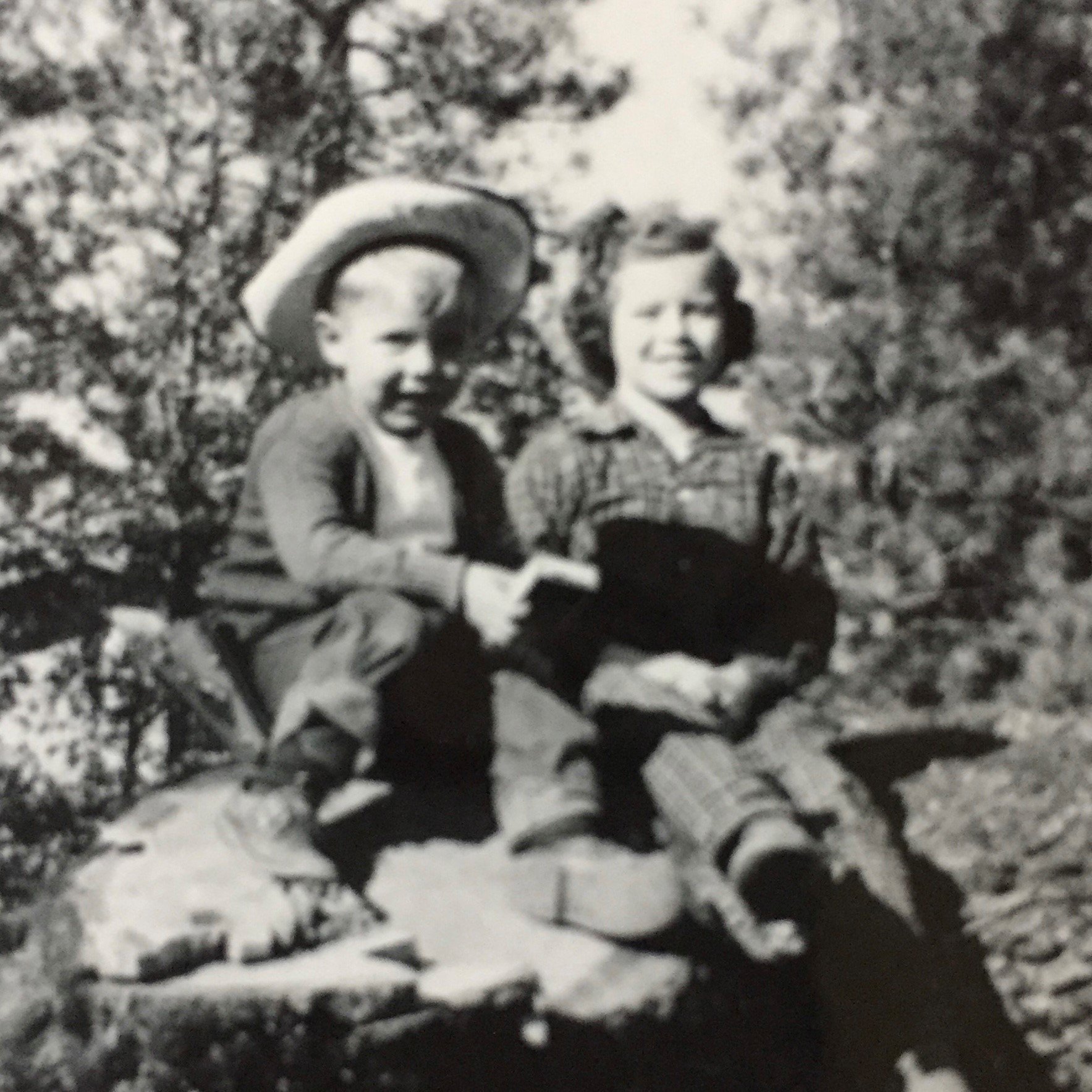Vintage black-and-white photograph of two young children, a boy with a wide-brimmed hat, sitting on a log.