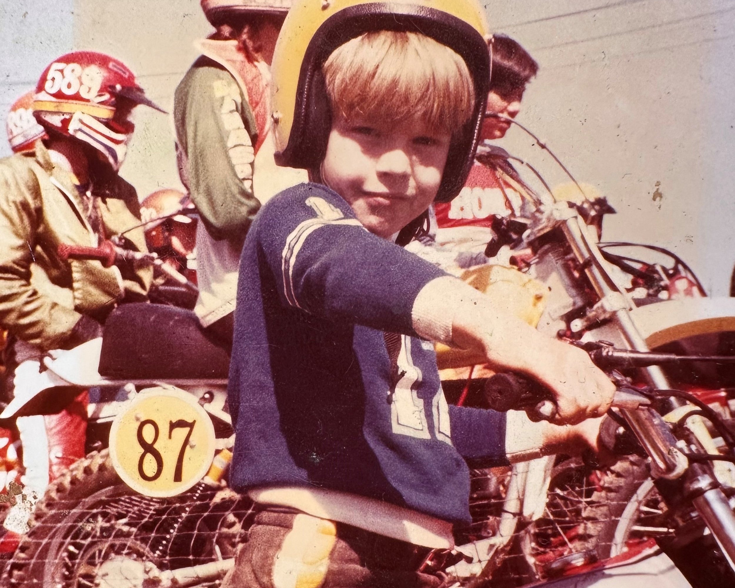 Vintage, faded photograph of a small child in a yellow helmet holding motorcycle handlebars, surrounded by other riders.