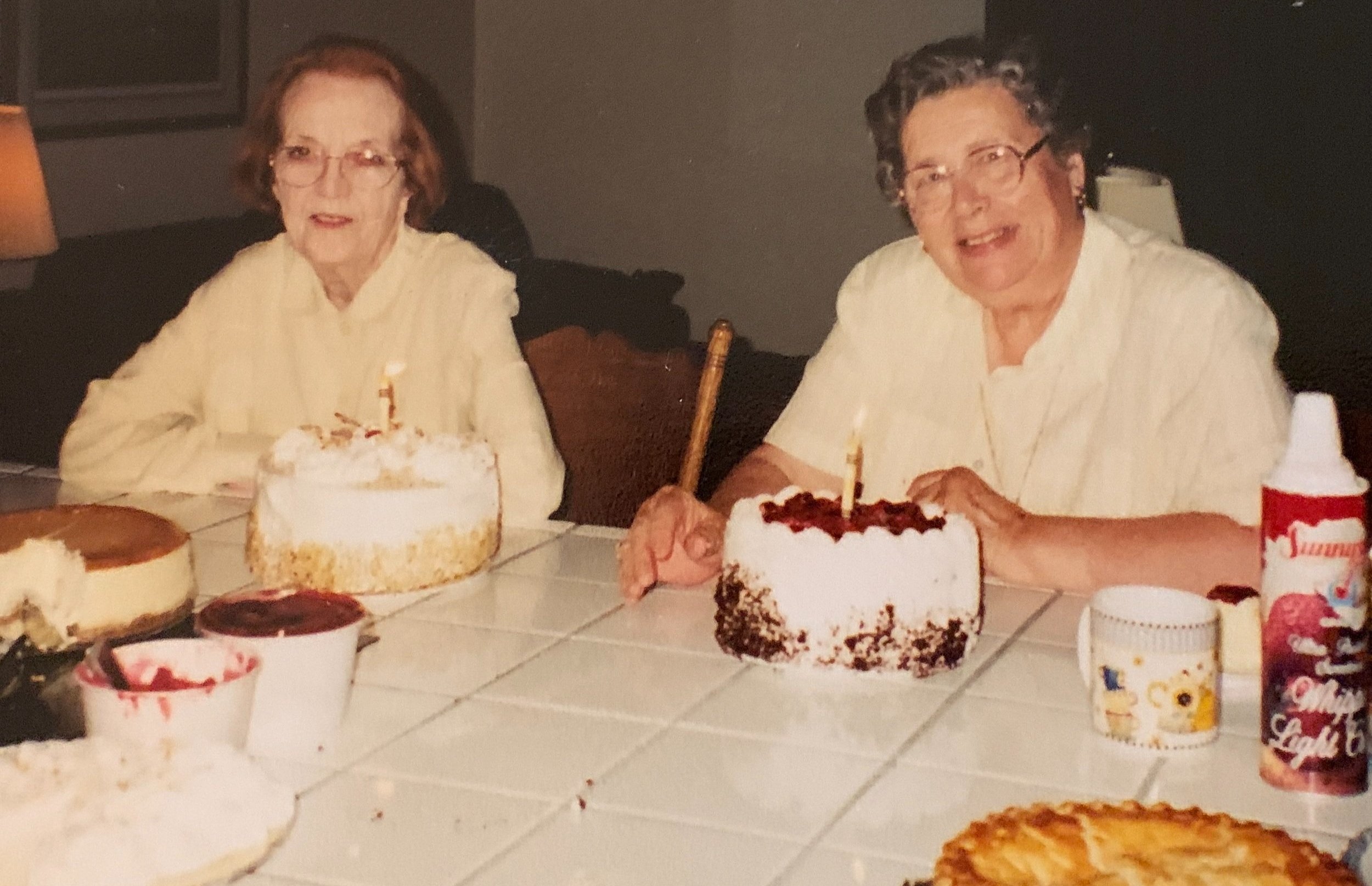 Two older women smiling behind a table with two birthday cakes and pies.