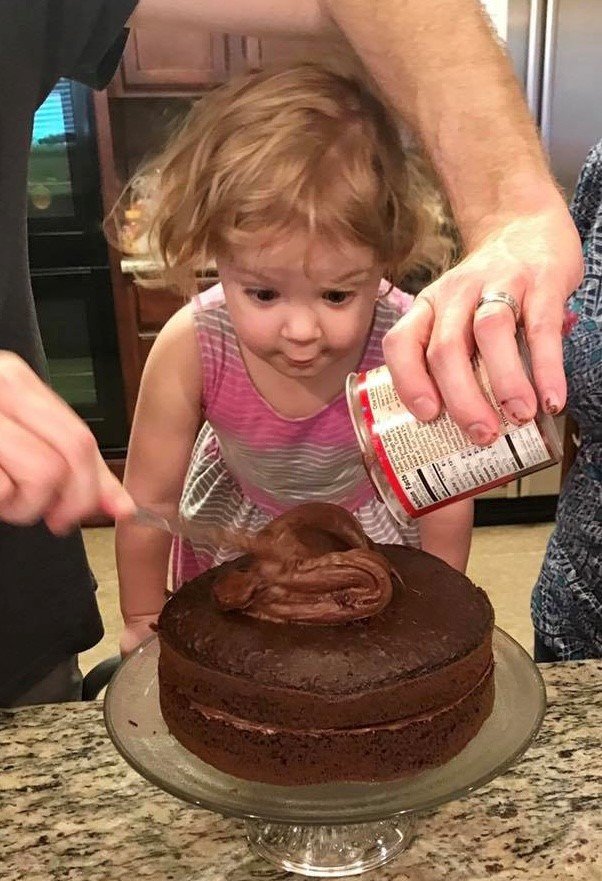 A young girl watching intently as an adult frosts a chocolate cake from a can.