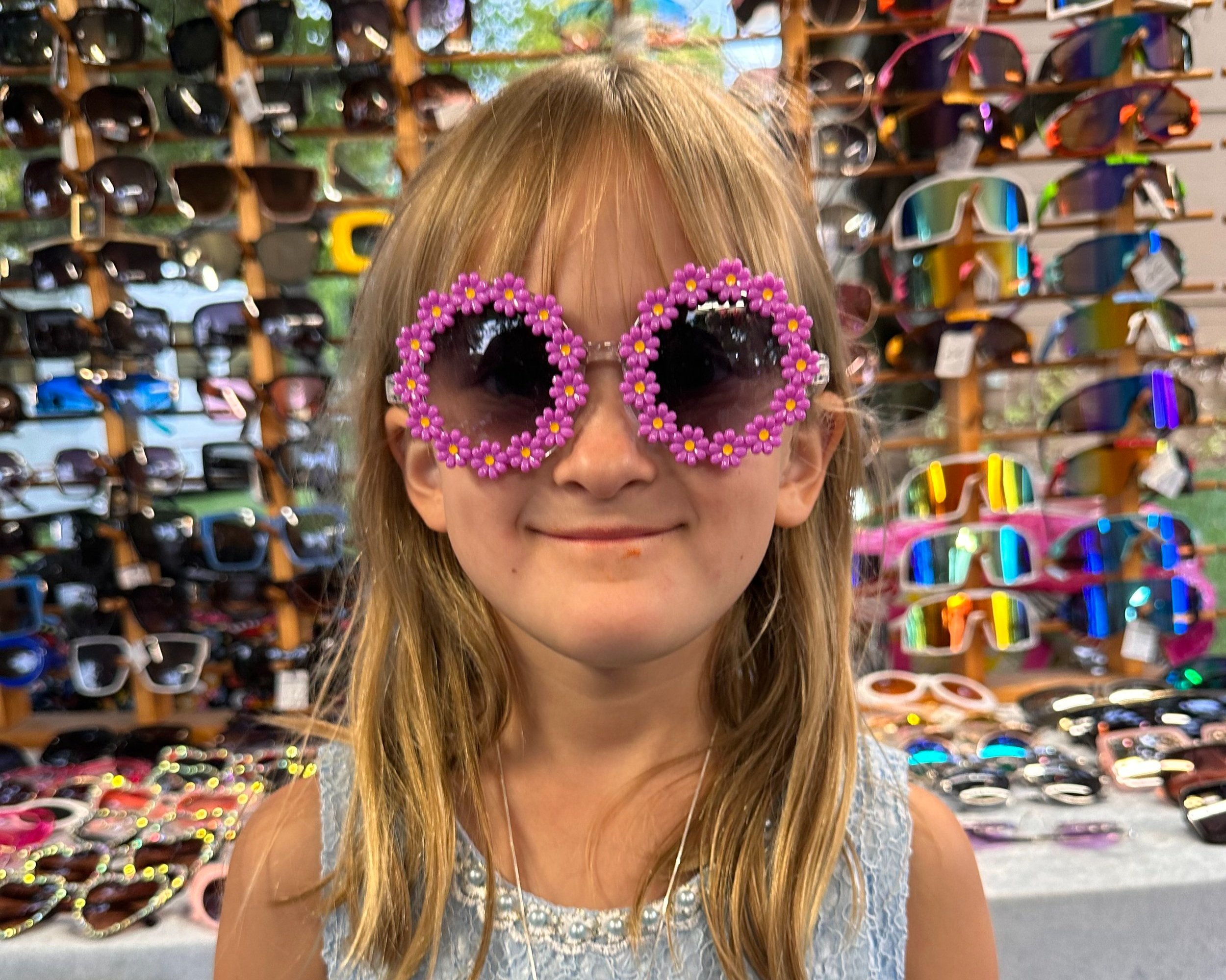 A young girl trying on large, playful sunglasses shaped like pink flowers.