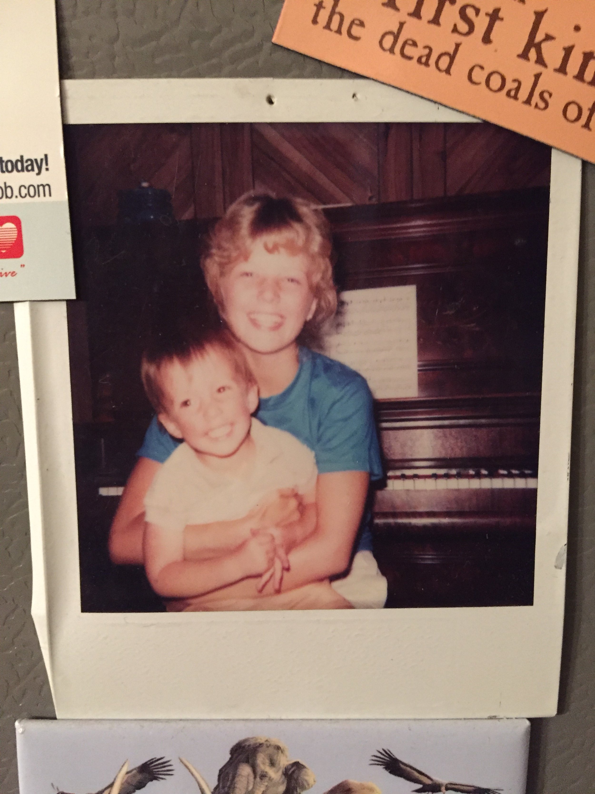 Vintage polaroid of an older girl smiling and hugging a younger boy, sitting in front of a piano.