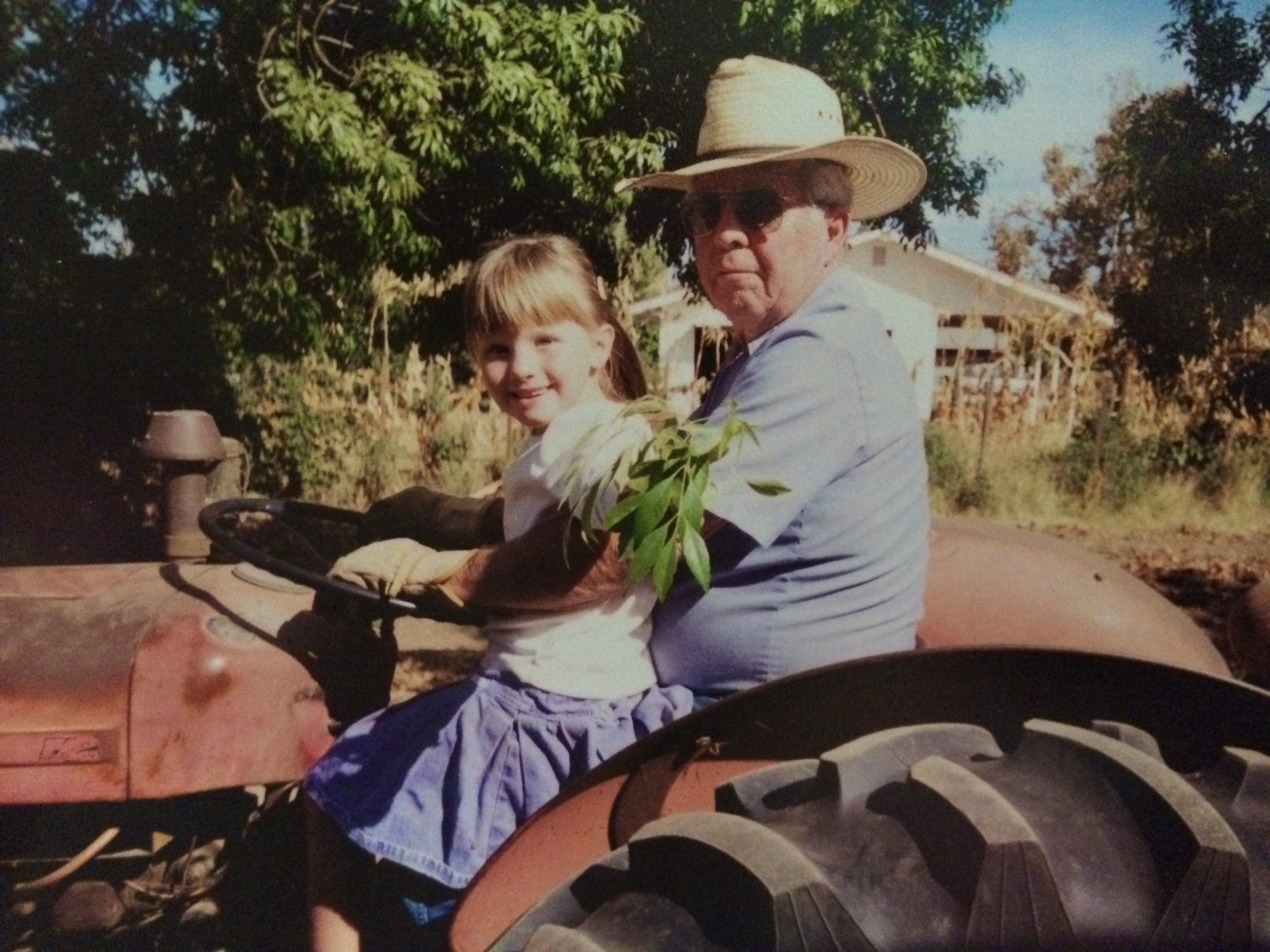 An older man wearing sunglasses and a straw hat driving a red tractor, with a young girl sitting on his lap.