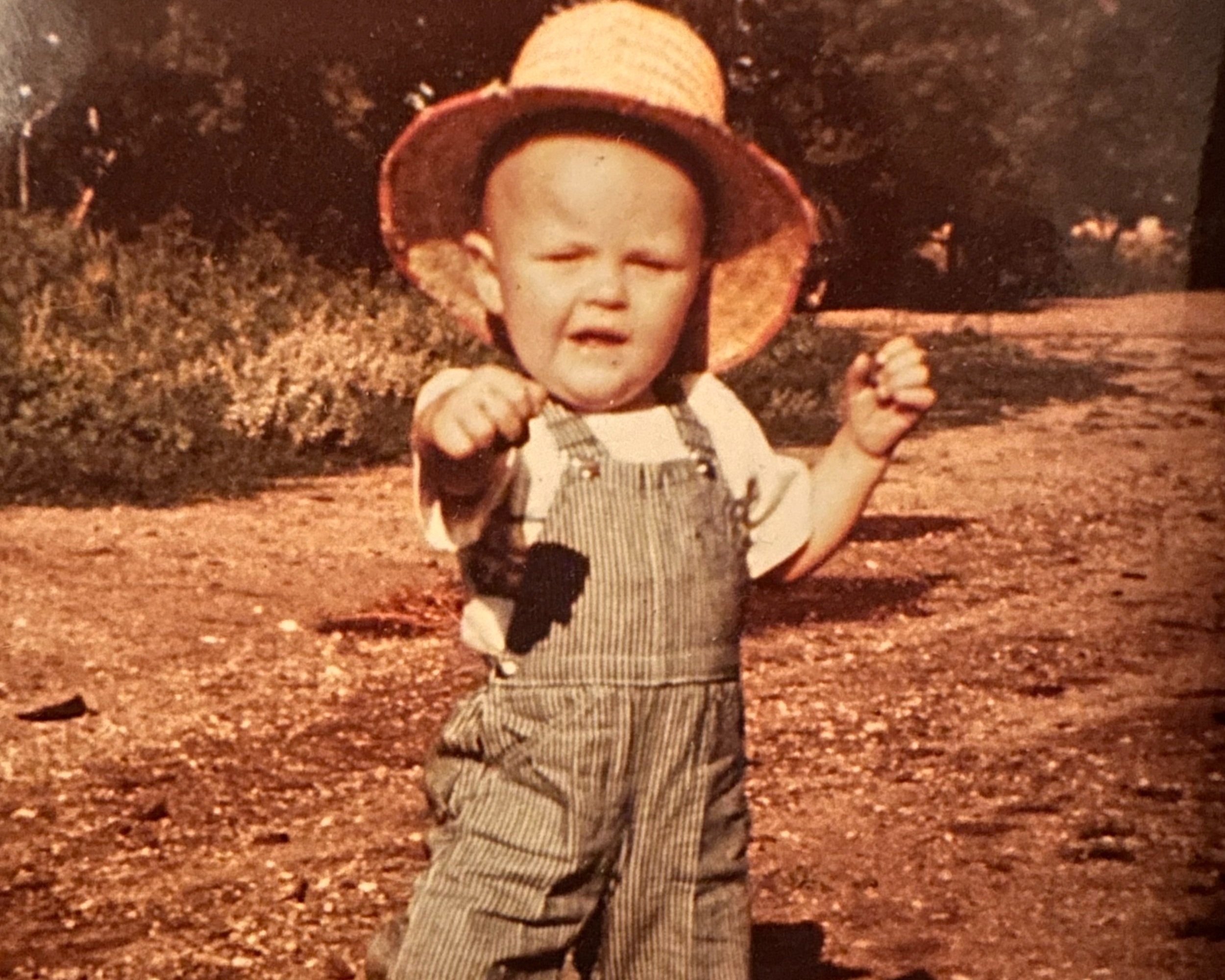 Vintage photo of a toddler boy in overalls and a straw hat looking tough at the camera.